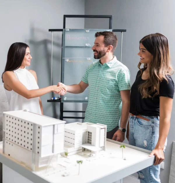 Architect and client shaking hands over a 3D white architectural building model in a professional design studio.