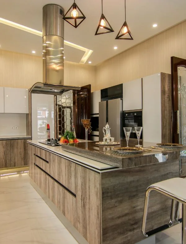 Modern modular kitchen design with a large wooden island, stainless steel ceiling-mounted chimney, and geometric pendant lights.