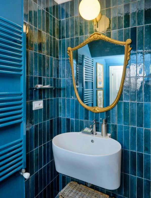 Vibrant bathroom featuring vertical teal blue subway tiles, a white wall-mounted sink, and a large ornate gold shield-shaped mirror.
