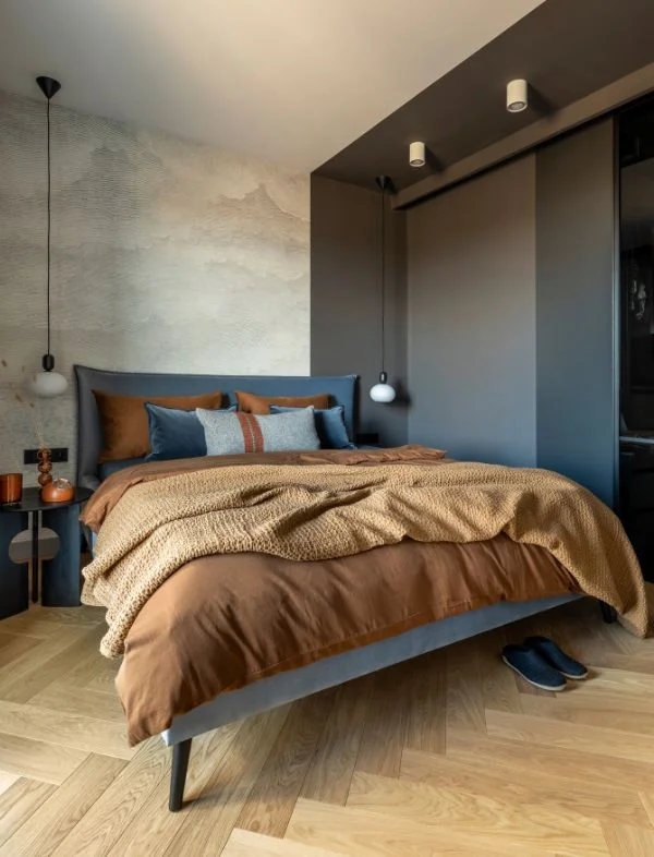 Modern bedroom interior with a grey upholstered bed, warm cinnamon brown bedding, herringbone wood flooring, and a sleek grey built-in wardrobe.