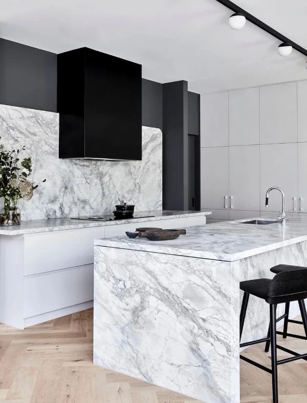 Modern luxury kitchen with a white marble waterfall island, matching marble backsplash, a matte black range hood, and light oak herringbone flooring.