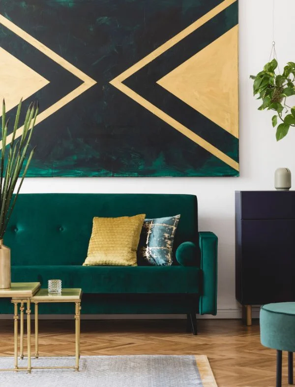 Modern living room featuring an emerald green velvet sofa with gold accent pillows, set against a large geometric teal and gold abstract painting on a white wall.