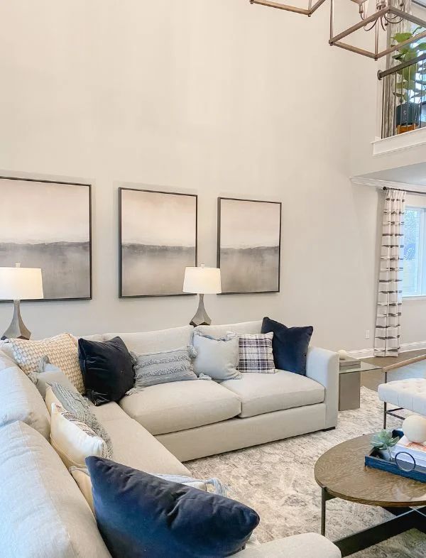 Modern transitional living room featuring a large light grey sectional sofa, a set of three abstract landscape wall art panels, and matching grey table lamps.