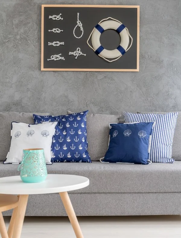 Modern nautical living room featuring a grey sofa with navy anchor and seashell patterned pillows, set against a textured grey concrete-effect wall with a framed lifebuoy and knot art piece.