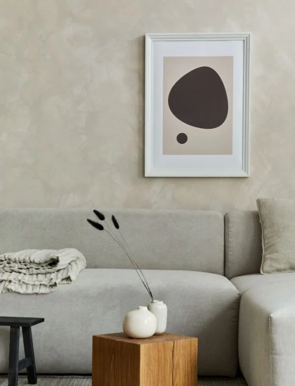 Modern living room featuring a beige limewash textured wall, a light grey minimalist sofa, and a framed abstract pebble art print in dark brown and tan.