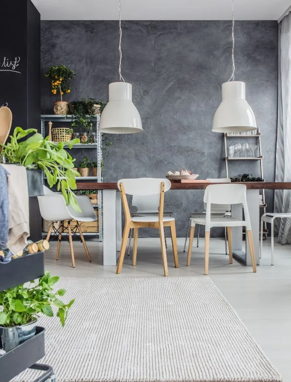 Modern industrial dining room featuring a dark grey textured plaster wall, a long wooden dining table with white designer chairs, and dual oversized white pendant lights.